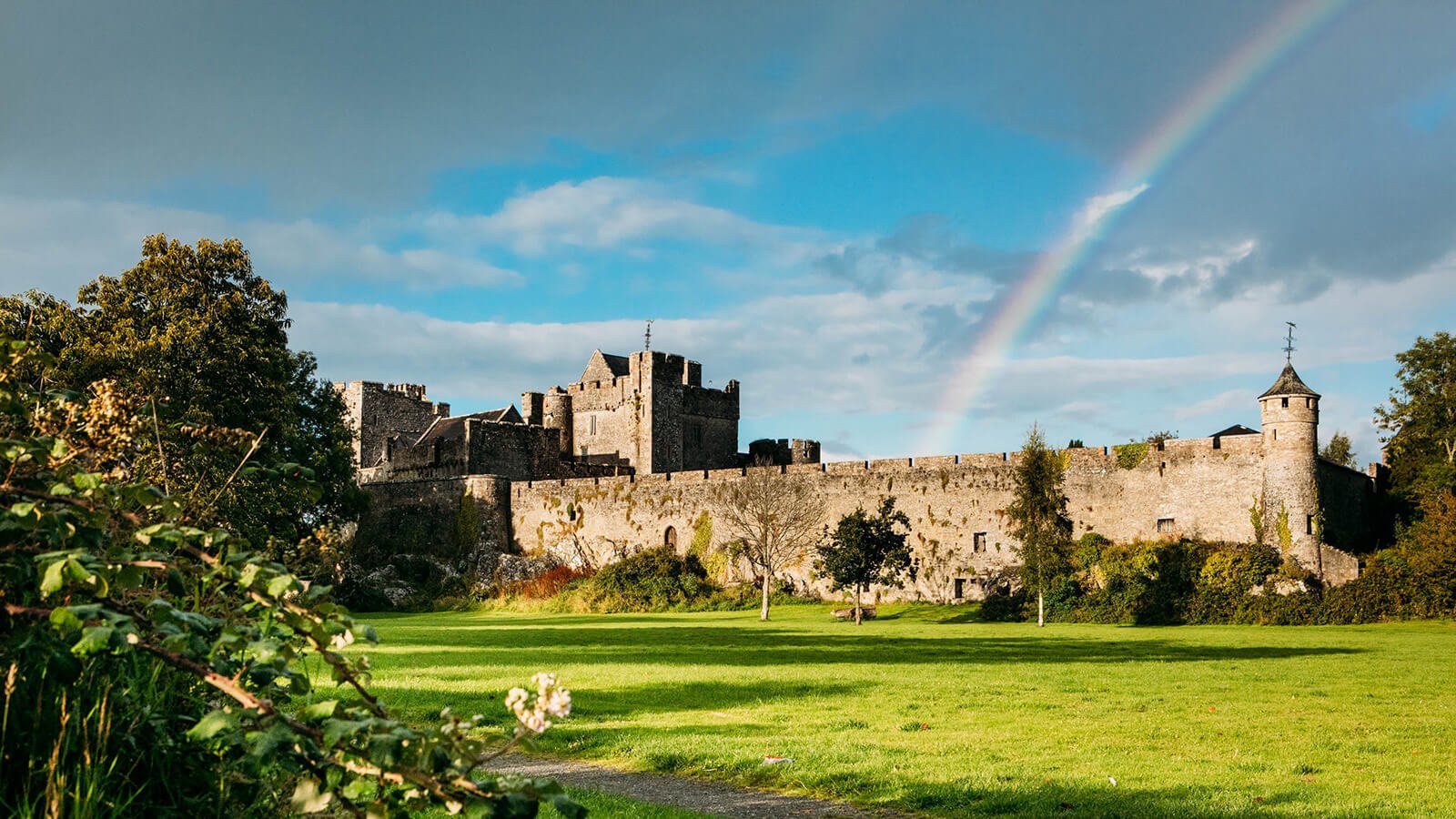 A rainbow over Cahir Castle walls with sunlit foliage in the foreground