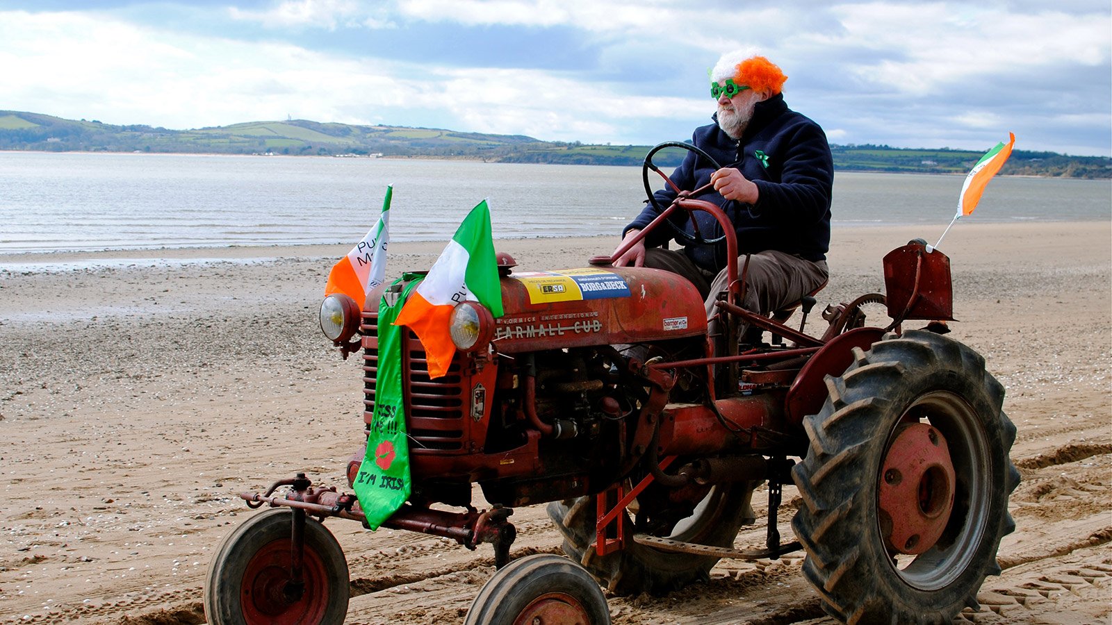 A stout man in a comedy wig and glasses driving a vintage tractor along a beach in Ireland