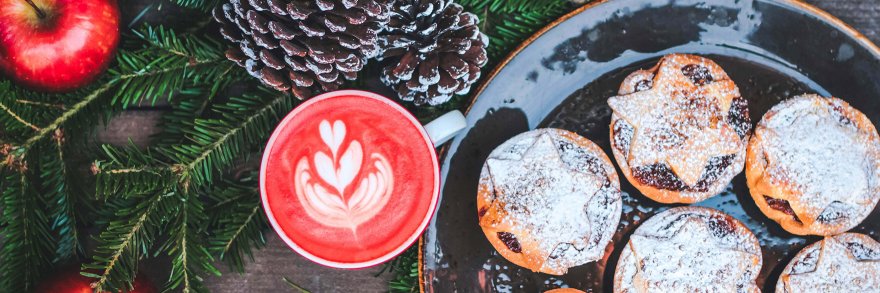 A plate of Christmas mince pies surrounded by pine cones, holly and a Christmas decorated coffee