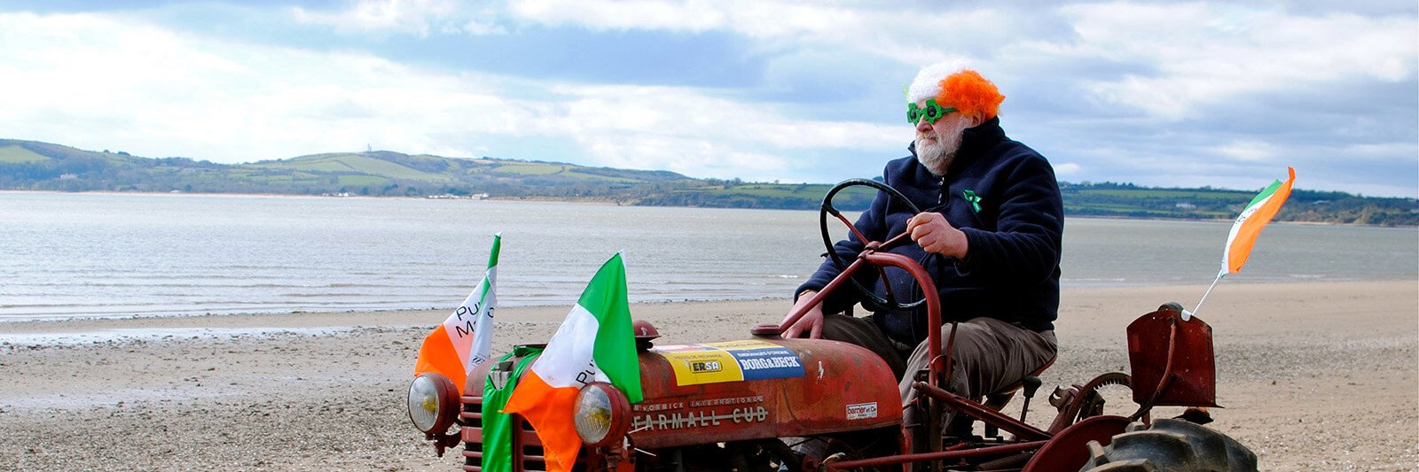 A man driving a tractor covered in Irish flags on a beach with an Irish wig and glasses on