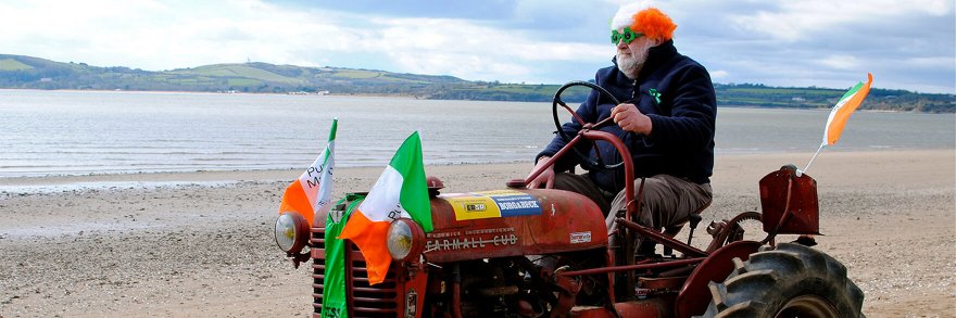 A farmer driving a tractor on a beach covered with Irish flags and an Irish wig