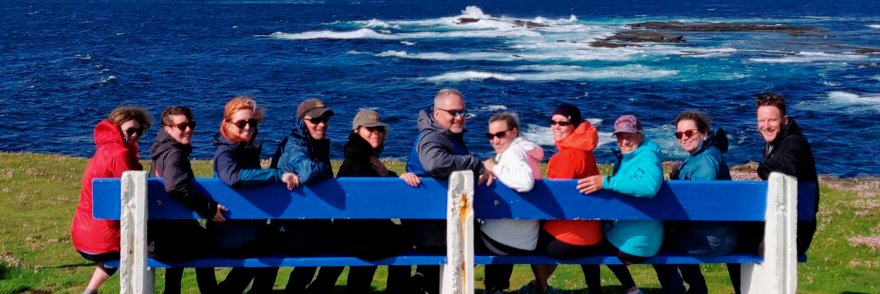 A Vagabond group smiling and seated on a blue bench in a scenic coastal location in Ireland