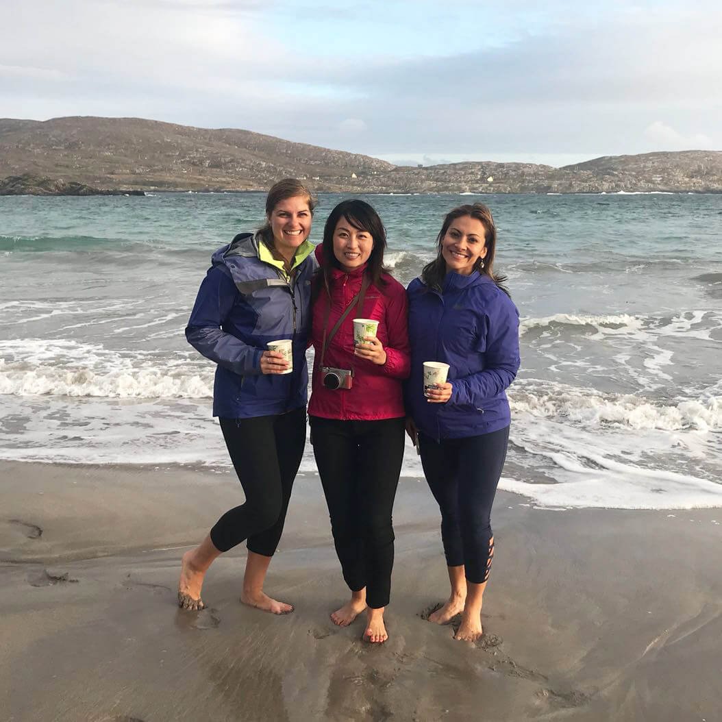 Three female Vagabond guests enjoy a wild brewed Irish coffee while standing barefoot together in the surf on beach
