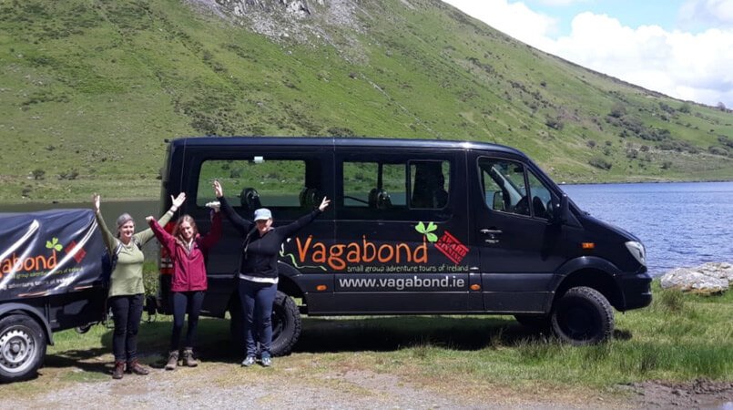 Three female Vagabond tour guests with their arms in the air in front of a VagaTron tour vehicle beside a lake and mountain