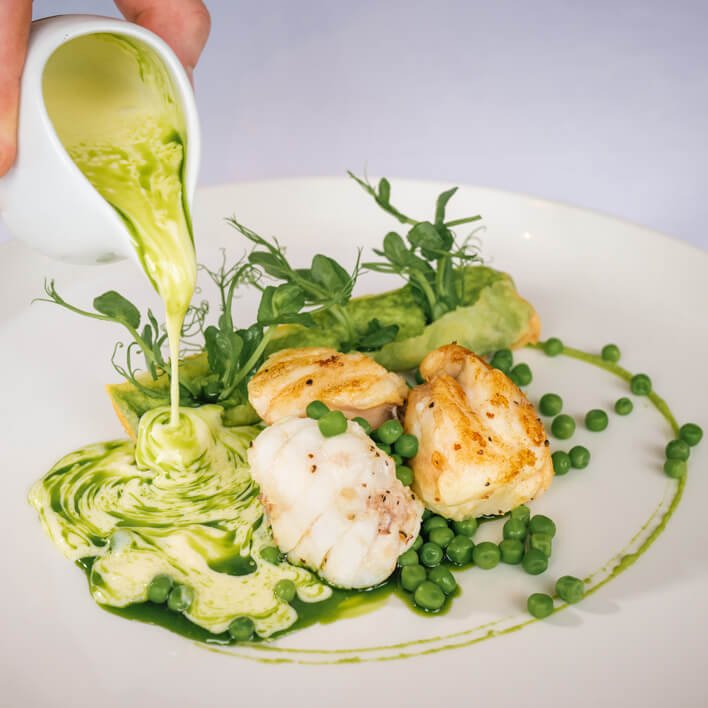 Hand pouring green sauce on monkfish medallions with peas