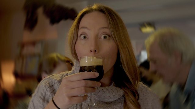 Female tour guest raises her eyebrows and widens her eyes at the camera while sipping from an Irish coffee