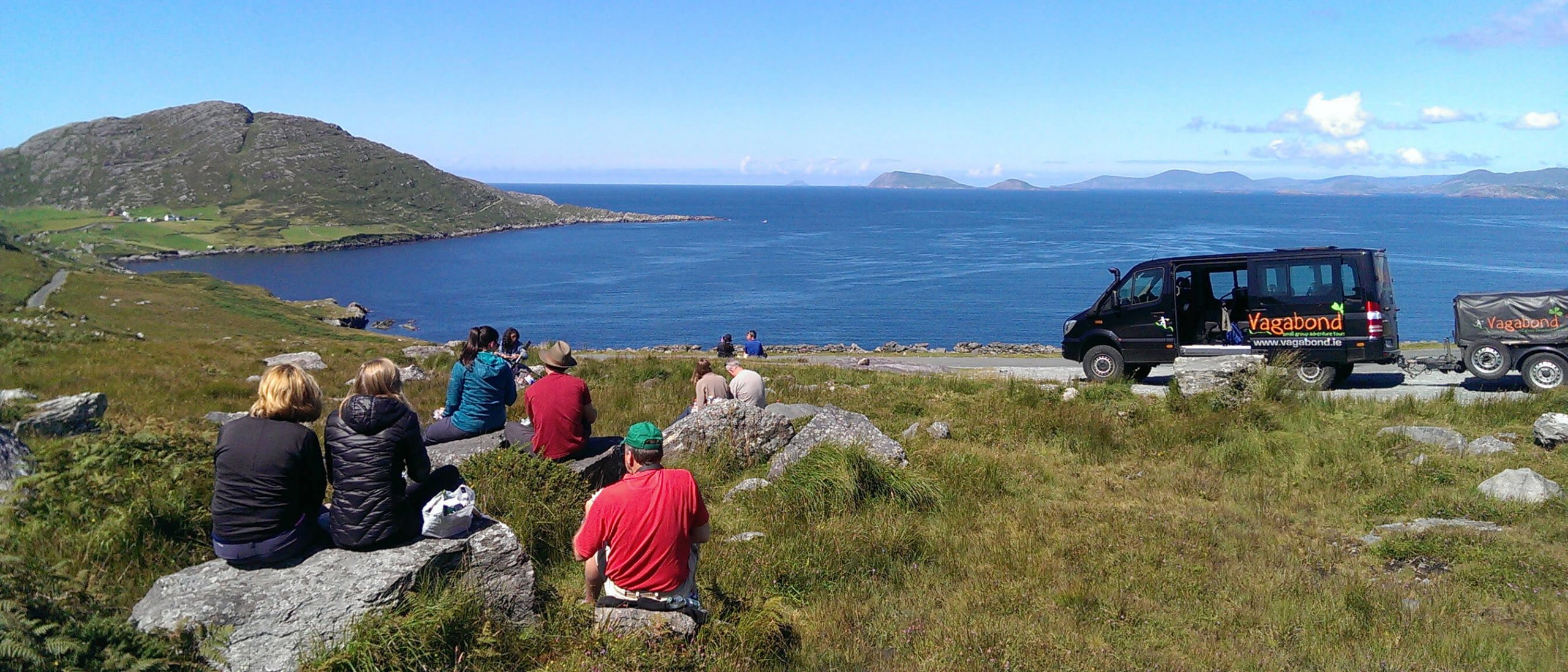 Vagabond group picnicing outside on a fine day with a VagaTron tour vehicle parked nearby and a scenic ocean view in the background