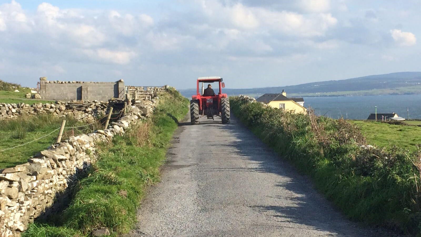 An empty, rural Irish laneway with a dry stone wall and red tractor driving into the distance