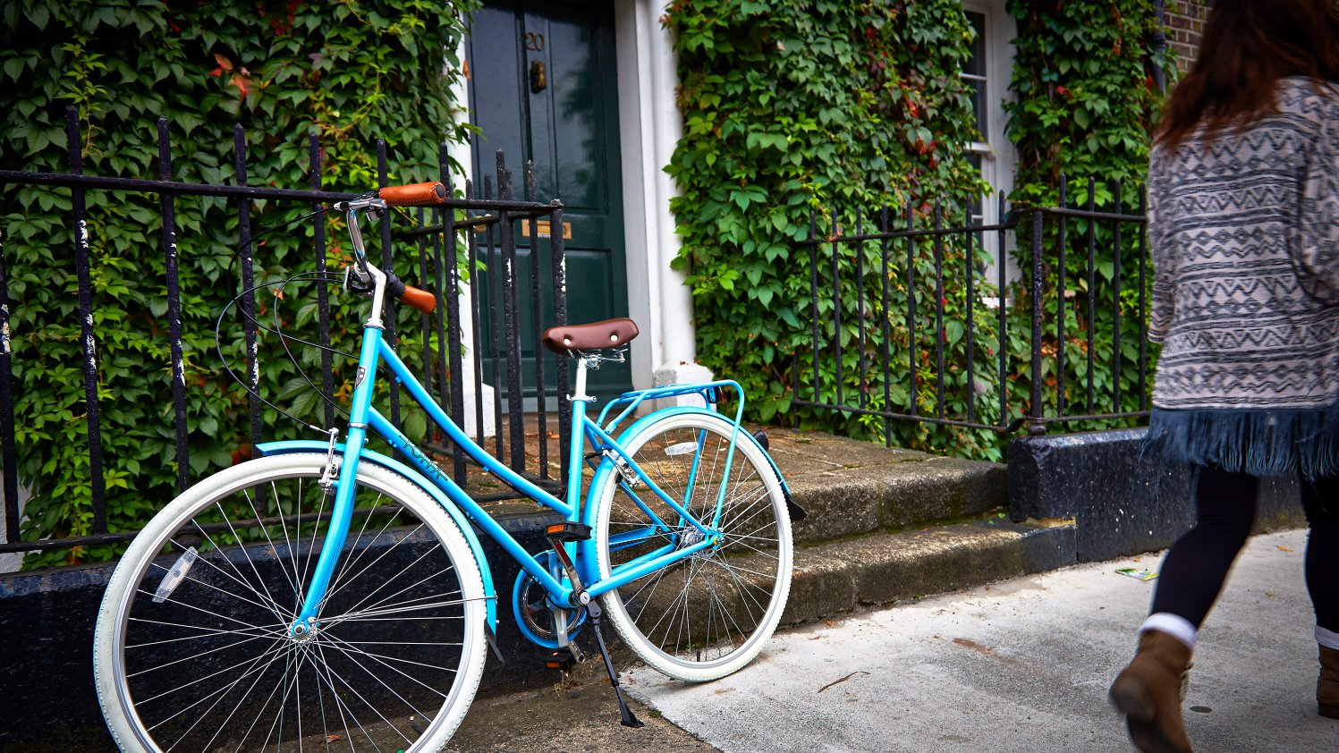A vibrantly blue bike leans against the railing of an ivy-coloured Georgian front door as a female walks by