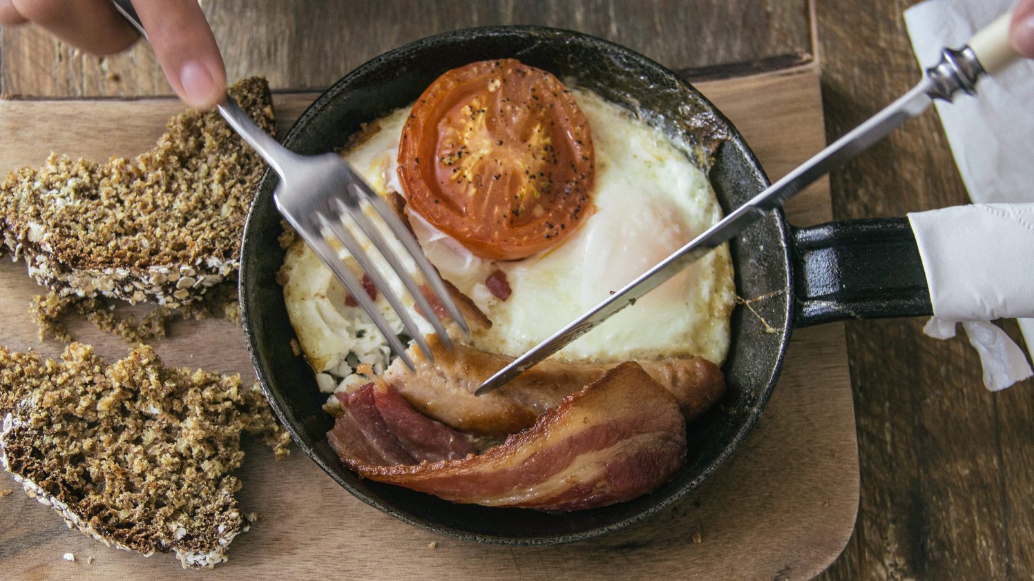 Hands holding knife and fork about to tuck into a cooked Irish breakfast, served in a frying pan